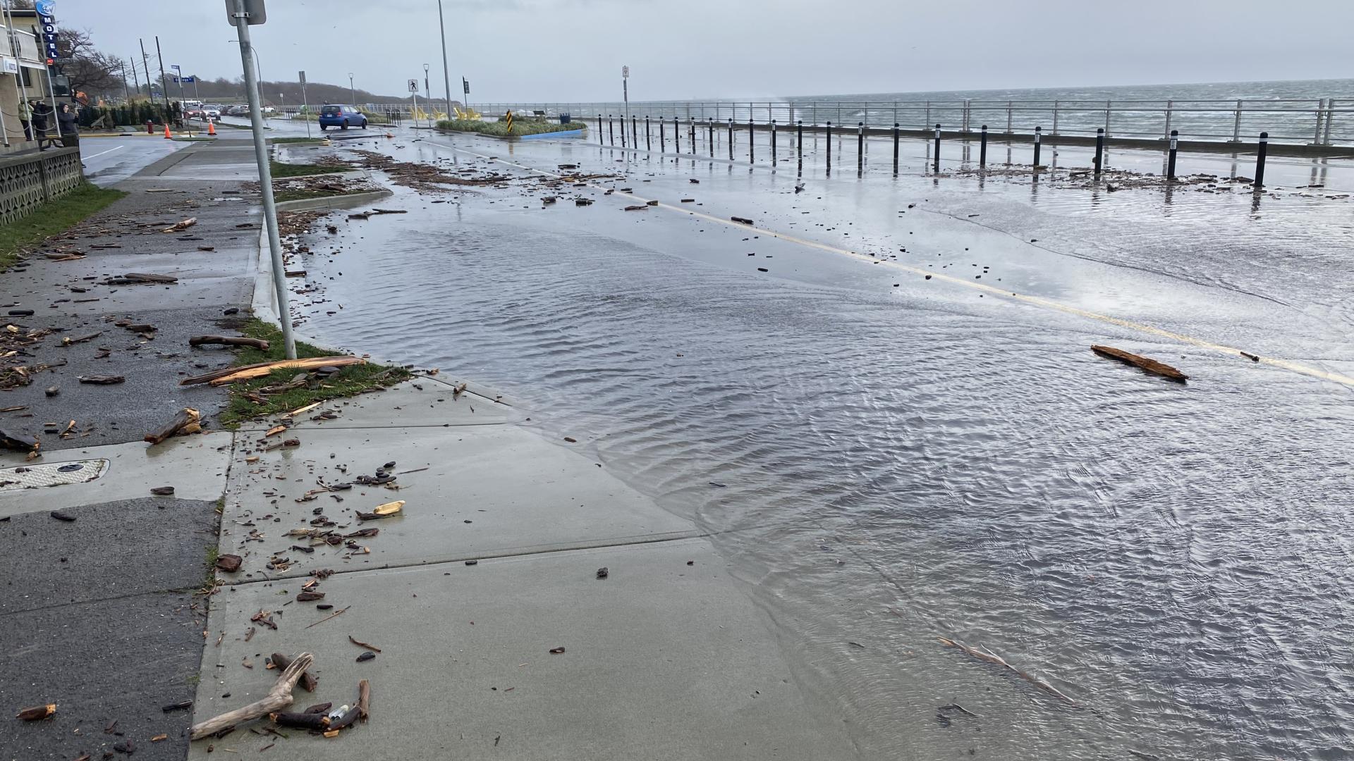 A picture of Dallas Road. The road, sidewalk and waterfront pathway in the background are flooded with water and covered in debris during a winter storm.